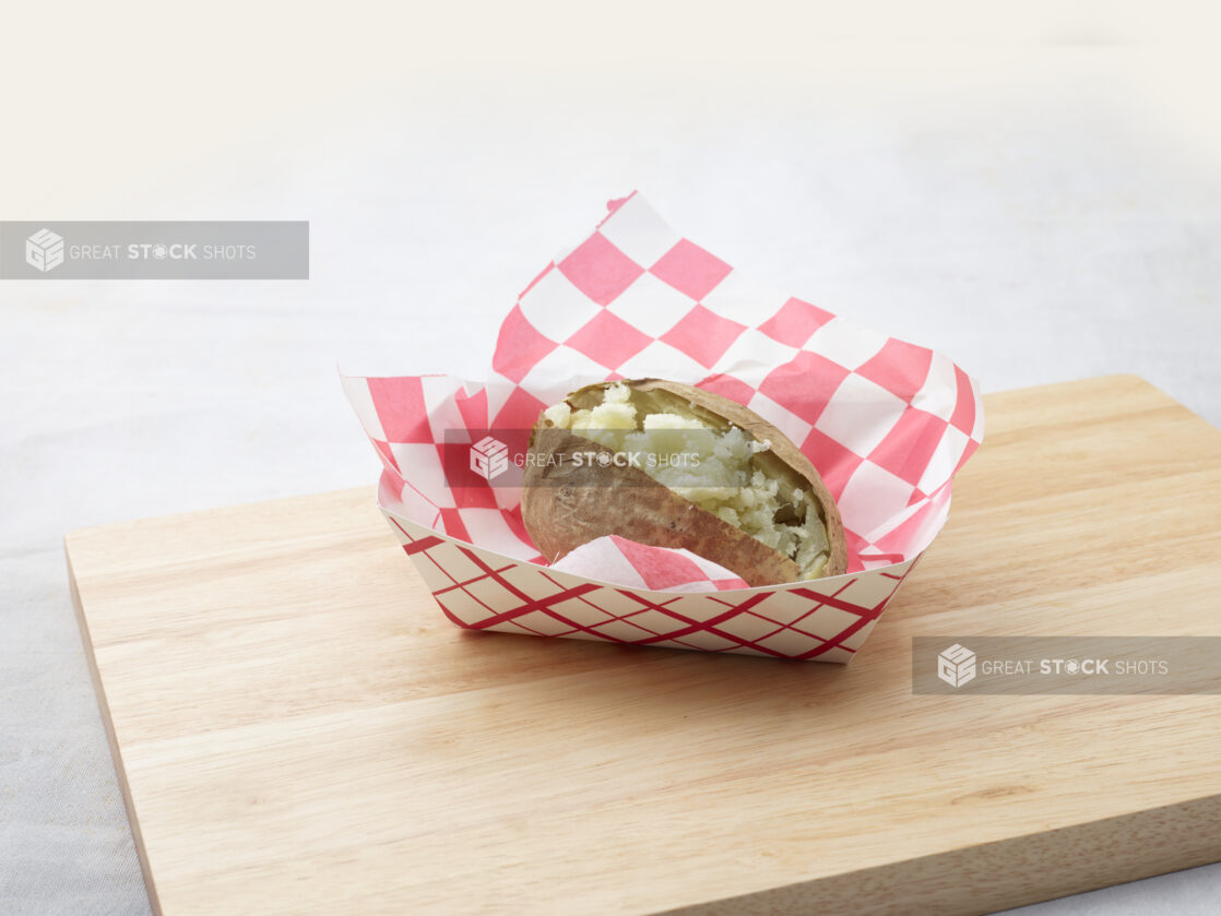 Split plain baked potato in a red and white lined cardboard takeout container on a wood board, white background