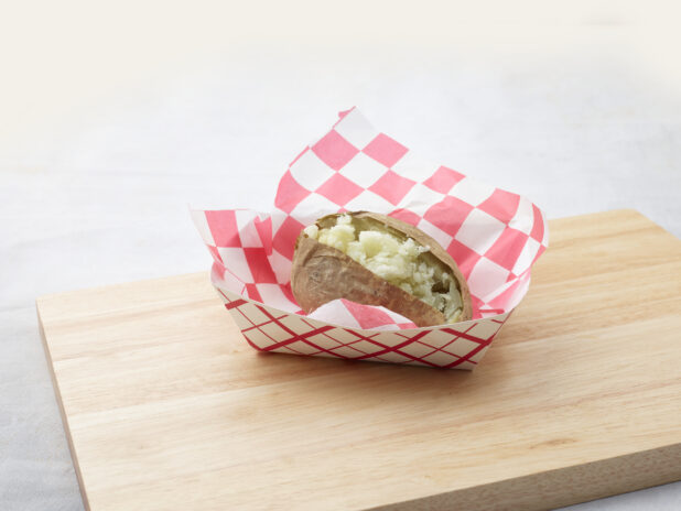 Split plain baked potato in a red and white lined cardboard takeout container on a wood board, white background