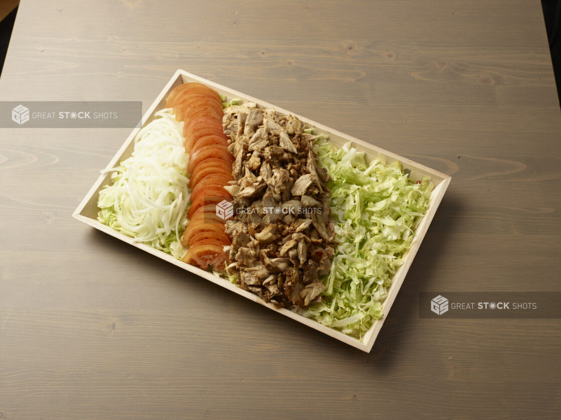 Platter of self-serve shawarma ingredients - chicken shawarma meat, shredded lettuce, sliced tomato, and sliced white onion on a wood catering tray, wood grain background