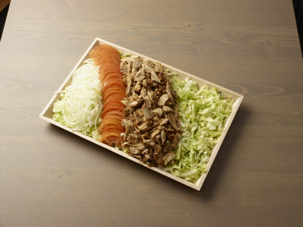 Platter of self-serve shawarma ingredients - chicken shawarma meat, shredded lettuce, sliced tomato, and sliced white onion on a wood catering tray, wood grain background