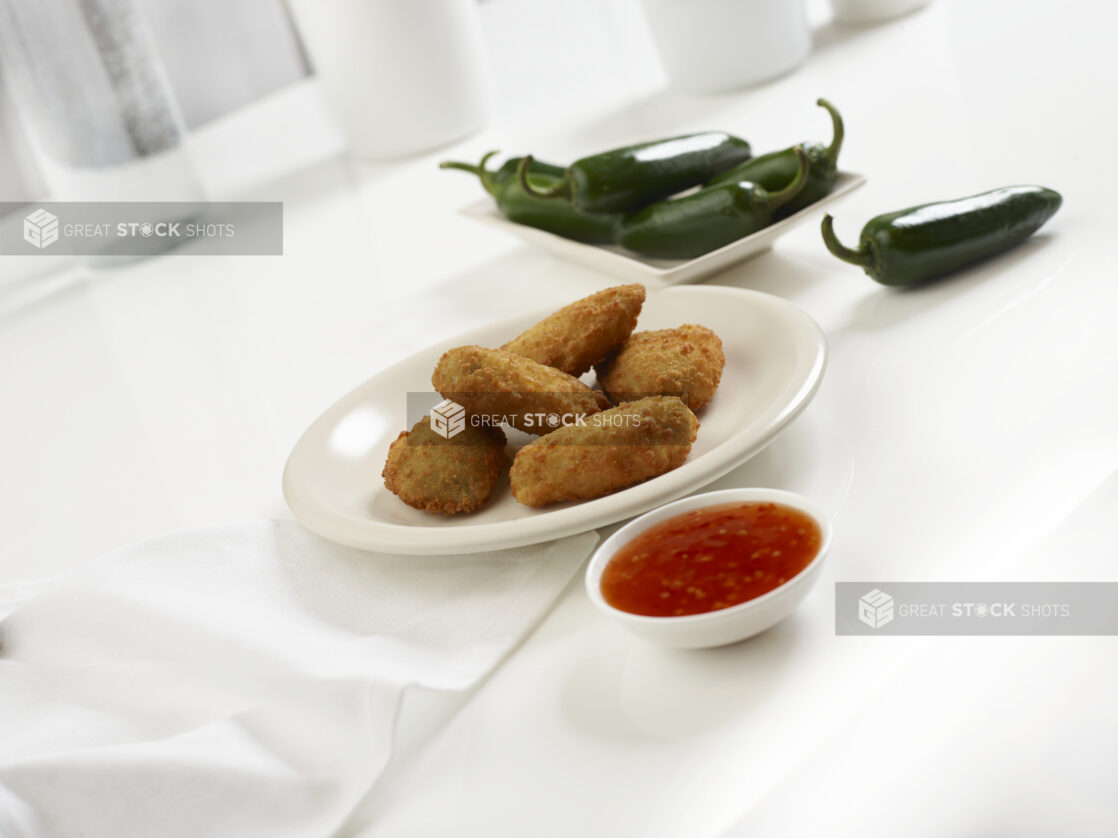 Five jalapeno poppers on a round white plate with chilli dipping sauce and whole jalapenos, white background