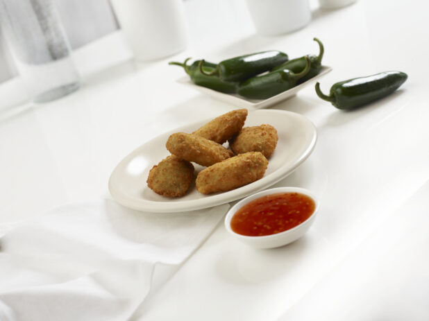 Five jalapeno poppers on a round white plate with chilli dipping sauce and whole jalapenos, white background