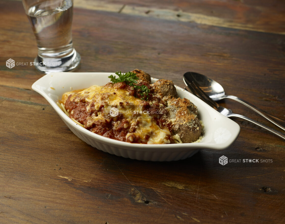 Broiled dish of spaghetti and meatballs topped with cheese and parsley on a wood tabletop