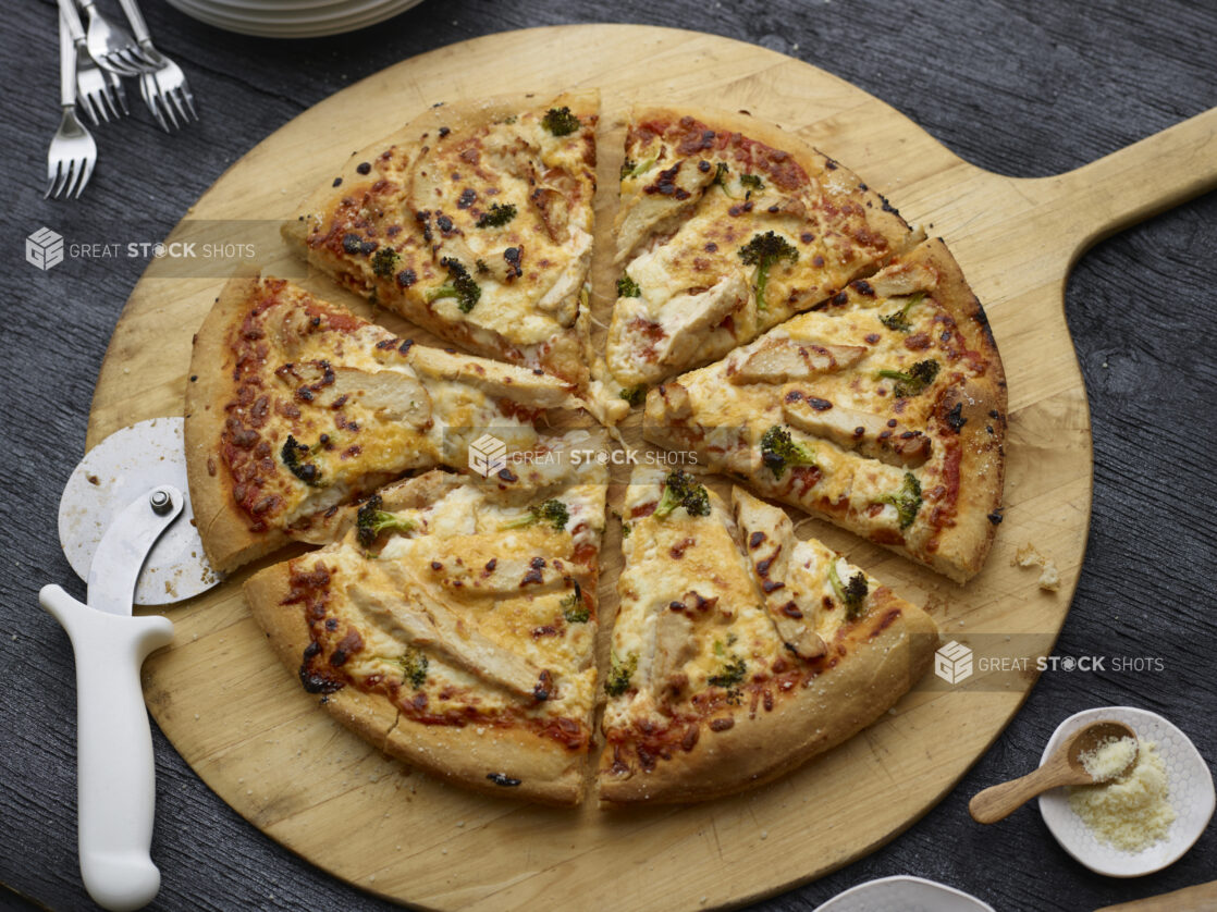 Whole sliced chicken, cheddar, and broccoli pizza on a round wood pizza peel with pizza cutter, grated parmesan with little wooden spoon in white dish alongside