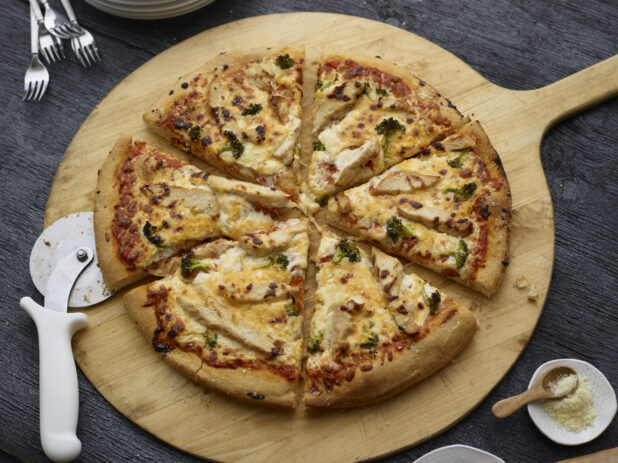 Whole sliced chicken, cheddar, and broccoli pizza on a round wood pizza peel with pizza cutter, grated parmesan with little wooden spoon in white dish alongside