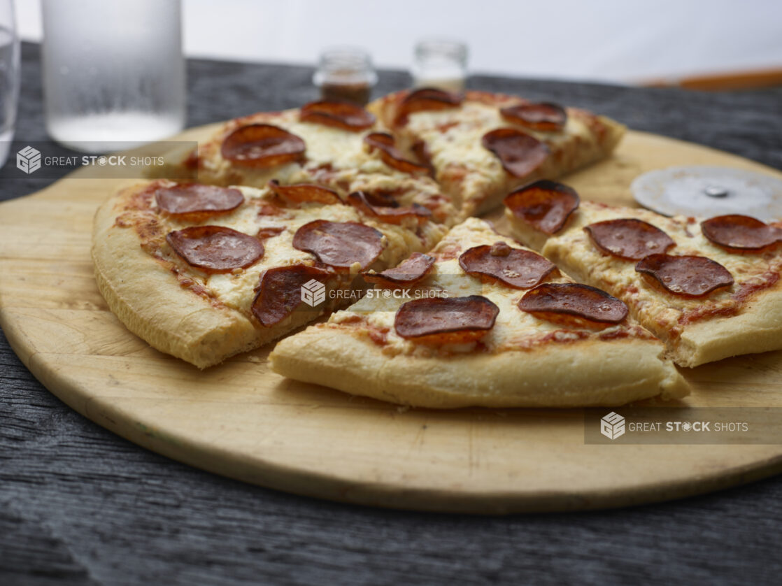 Whole sliced pepperoni pizza on a round wood pizza peel on a black tabletop