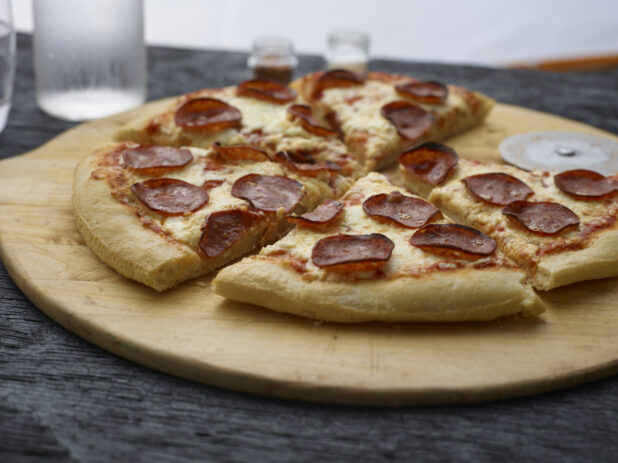 Whole sliced pepperoni pizza on a round wood pizza peel on a black tabletop