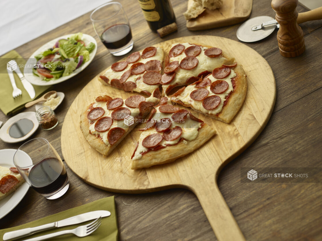 Sliced pepperoni pizza on a round wooden pizza peel with cutter, side salad, utensils, wine glasses in background