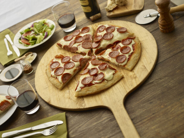 Sliced pepperoni pizza on a round wooden pizza peel with cutter, side salad, utensils, wine glasses in background