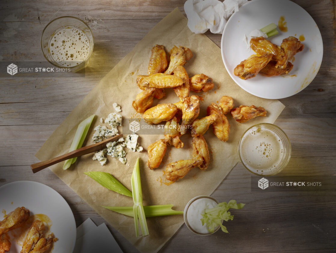 Sauced chicken wings on parchment paper with blue cheese and celery sticks, wings on sideplates and glasses of beer surrounding