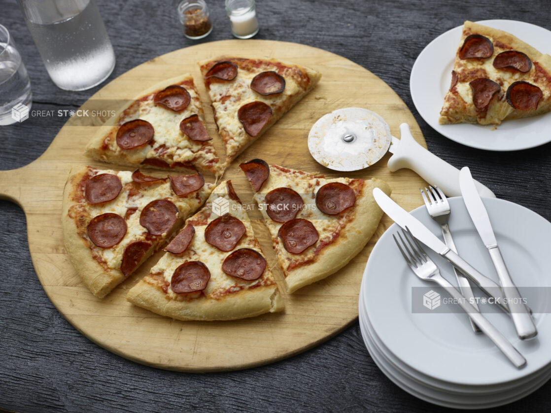 Sliced pepperoni pizza on a round wooden pizza peel with cutter, side plates, and cutlery, slice on plate