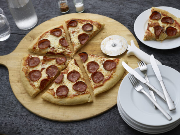Sliced pepperoni pizza on a round wooden pizza peel with cutter, side plates, and cutlery, slice on plate