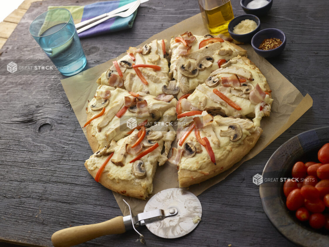 Whole sliced bacon, chicken, mushroom, and red pepper pizza with a cutter on parchment paper with utensils and condiments in the background