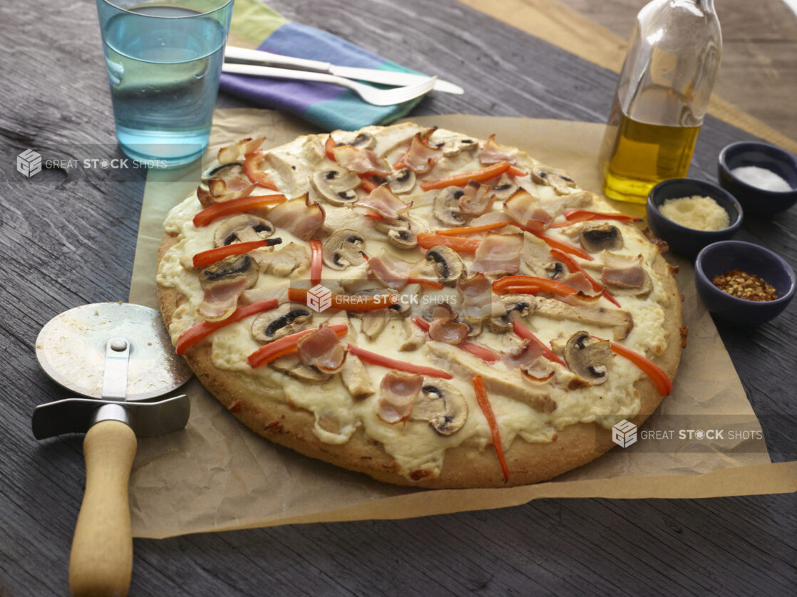 Whole unsliced bacon, chicken, mushroom, and red pepper pizza with a cutter on parchment paper with utensils and condiments in the background