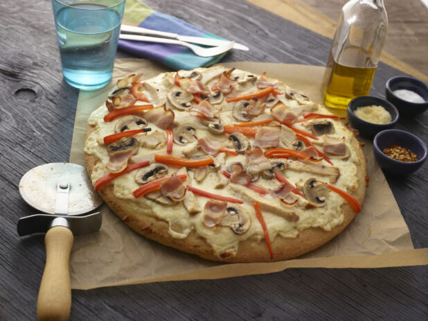 Whole unsliced bacon, chicken, mushroom, and red pepper pizza with a cutter on parchment paper with utensils and condiments in the background