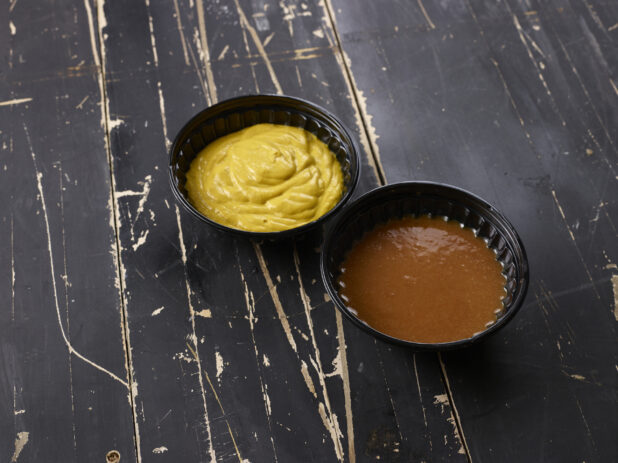 Yellow mustard and barbecue sauce in round black plastic ramekins on a weathered black-painted wood surface