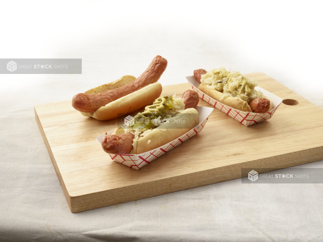 Three ballpark-style hot dogs, various toppings, arranged on a wood board, light canvas background