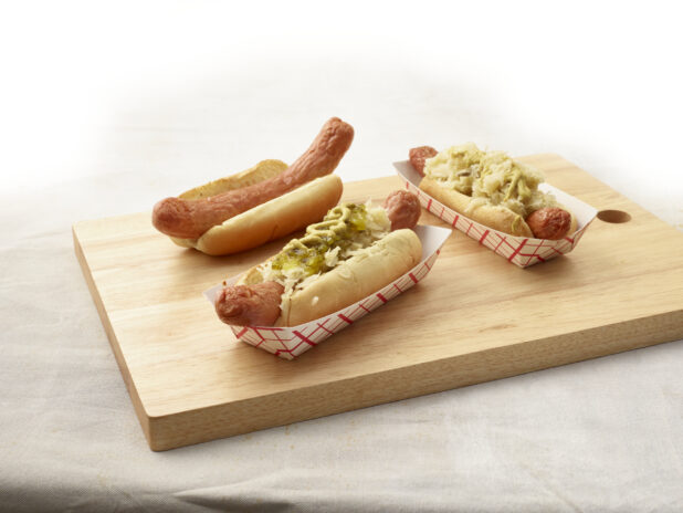 Three ballpark-style hot dogs, various toppings, arranged on a wood board, light canvas background