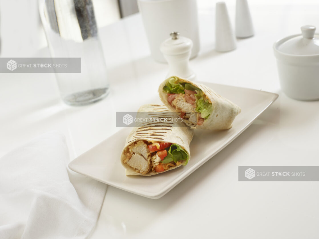 Fried chicken, bacon, lettuce, tomato, and cheddar wrap, halved, on a white plate, in an all-white table setting