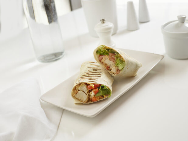 Fried chicken, bacon, lettuce, tomato, and cheddar wrap, halved, on a white plate, in an all-white table setting