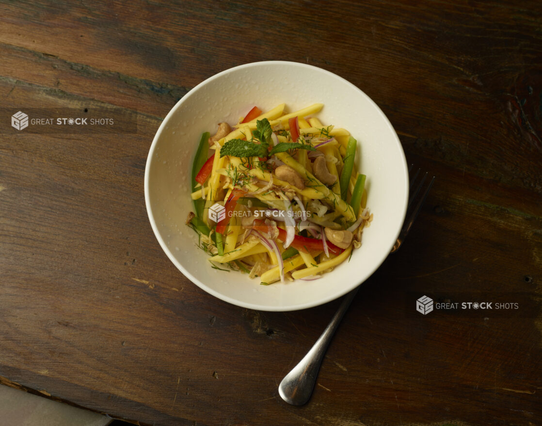 Mango salad with cashews, peppers, red onion, and herb garnish in a round white bowl with a fork on a dark wood background, overhead view