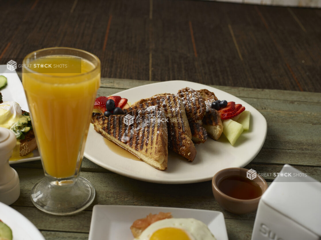 French toast with maple syrup and fresh fruit with a tall glass of orange juice on a table with other breakfast food surrounding