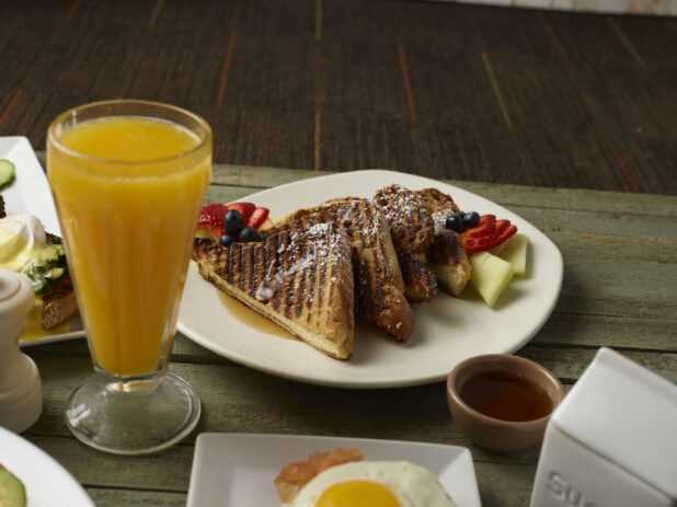 French toast with maple syrup and fresh fruit with a tall glass of orange juice on a table with other breakfast food surrounding