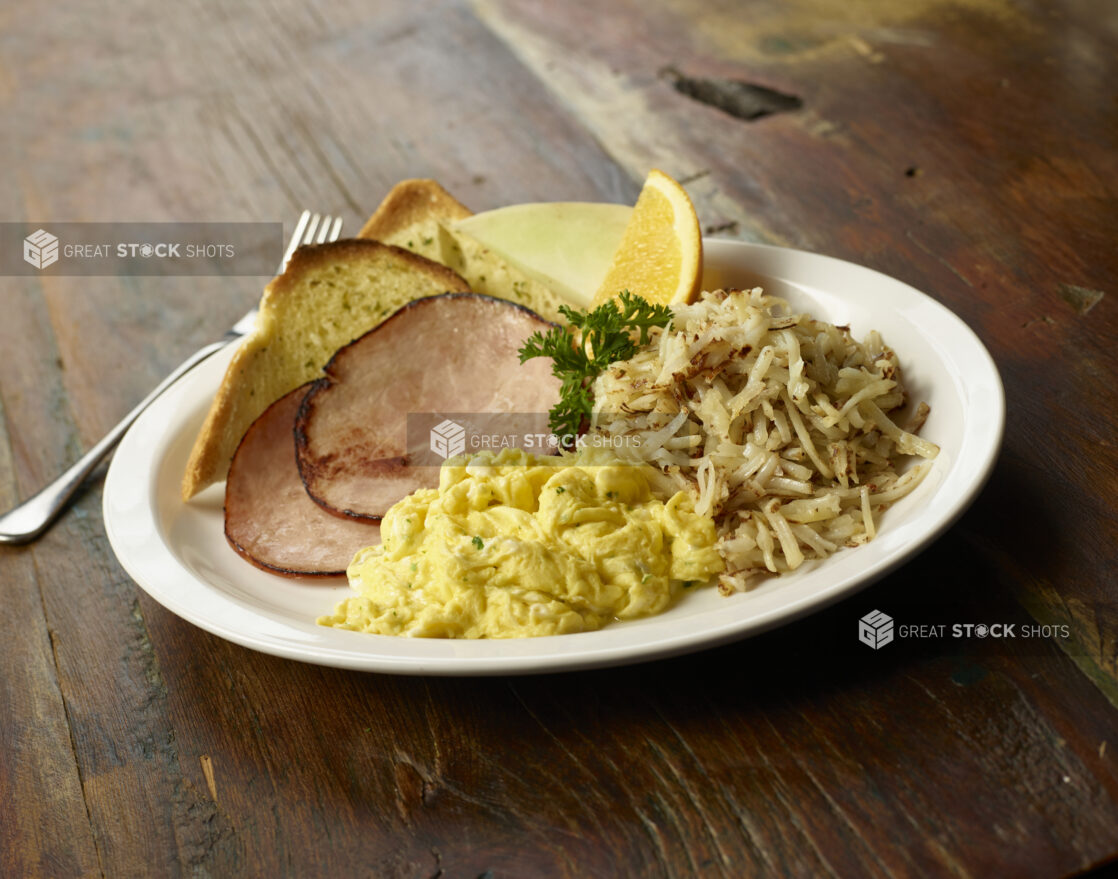 Scrambled eggs, fried ham, hash browns, and garlic toast on a white plate on a dark wood tabletop, close-up