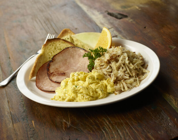 Scrambled eggs, fried ham, hash browns, and garlic toast on a white plate on a dark wood tabletop, close-up