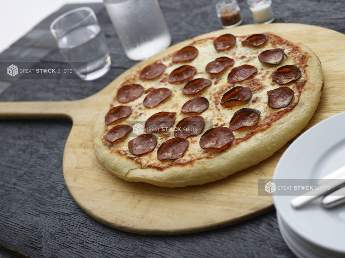 Whole pepperoni pizza on a wooden pizza peel with white plates, cutlery and water glasses