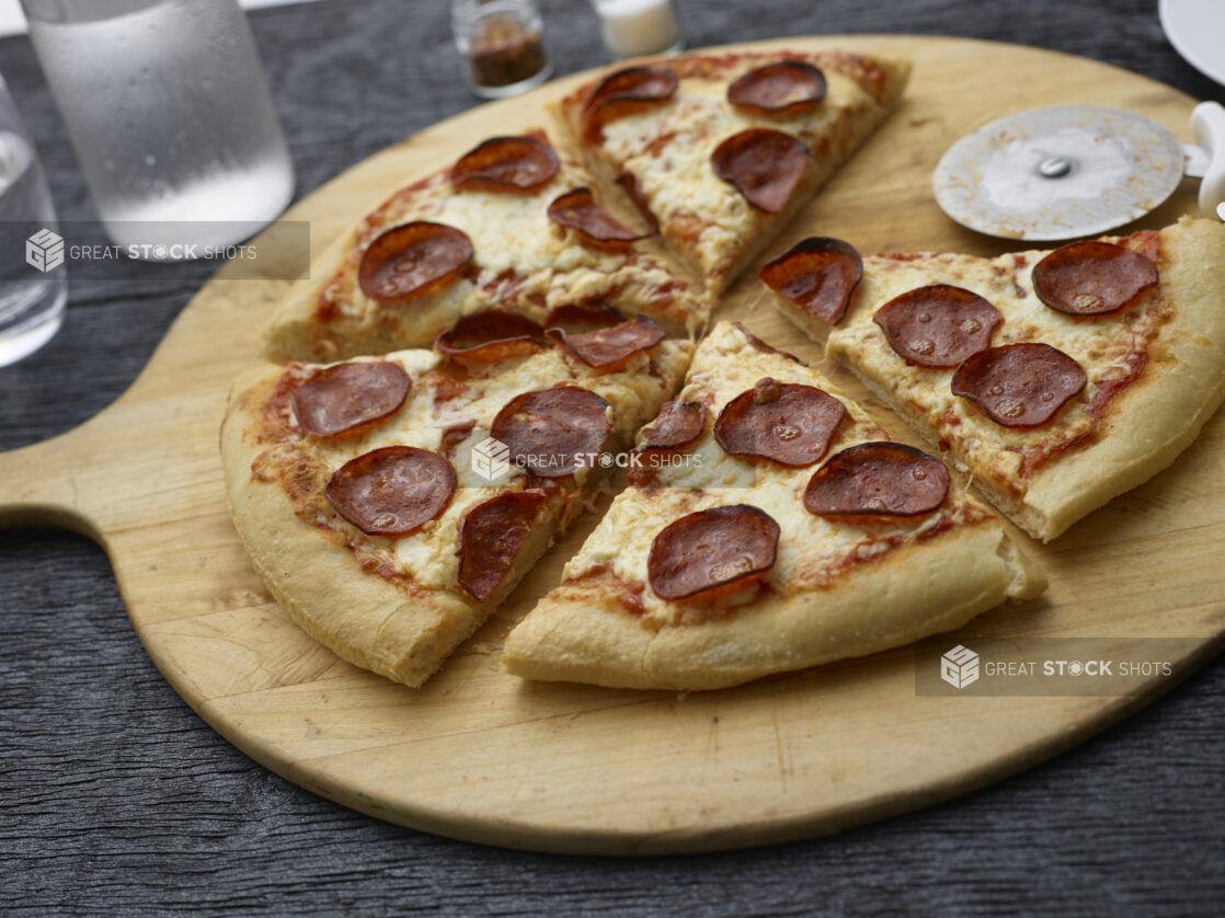 Sliced pepperoni pizza on a round wood pizza peel with pizza cutter, black background,