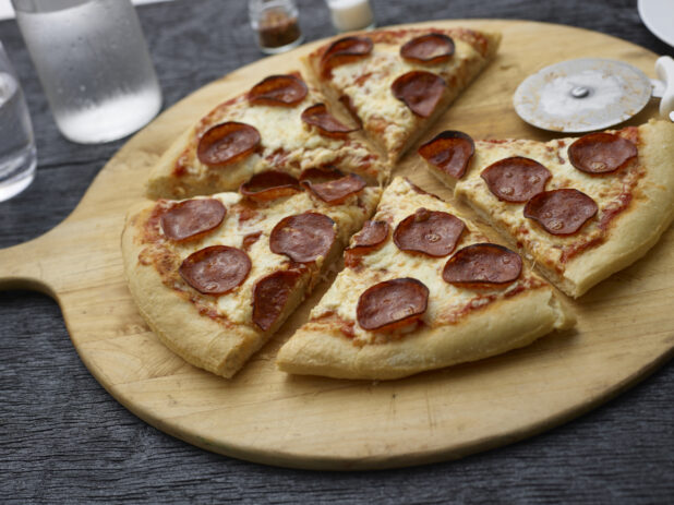 Sliced pepperoni pizza on a round wood pizza peel with pizza cutter, black background,