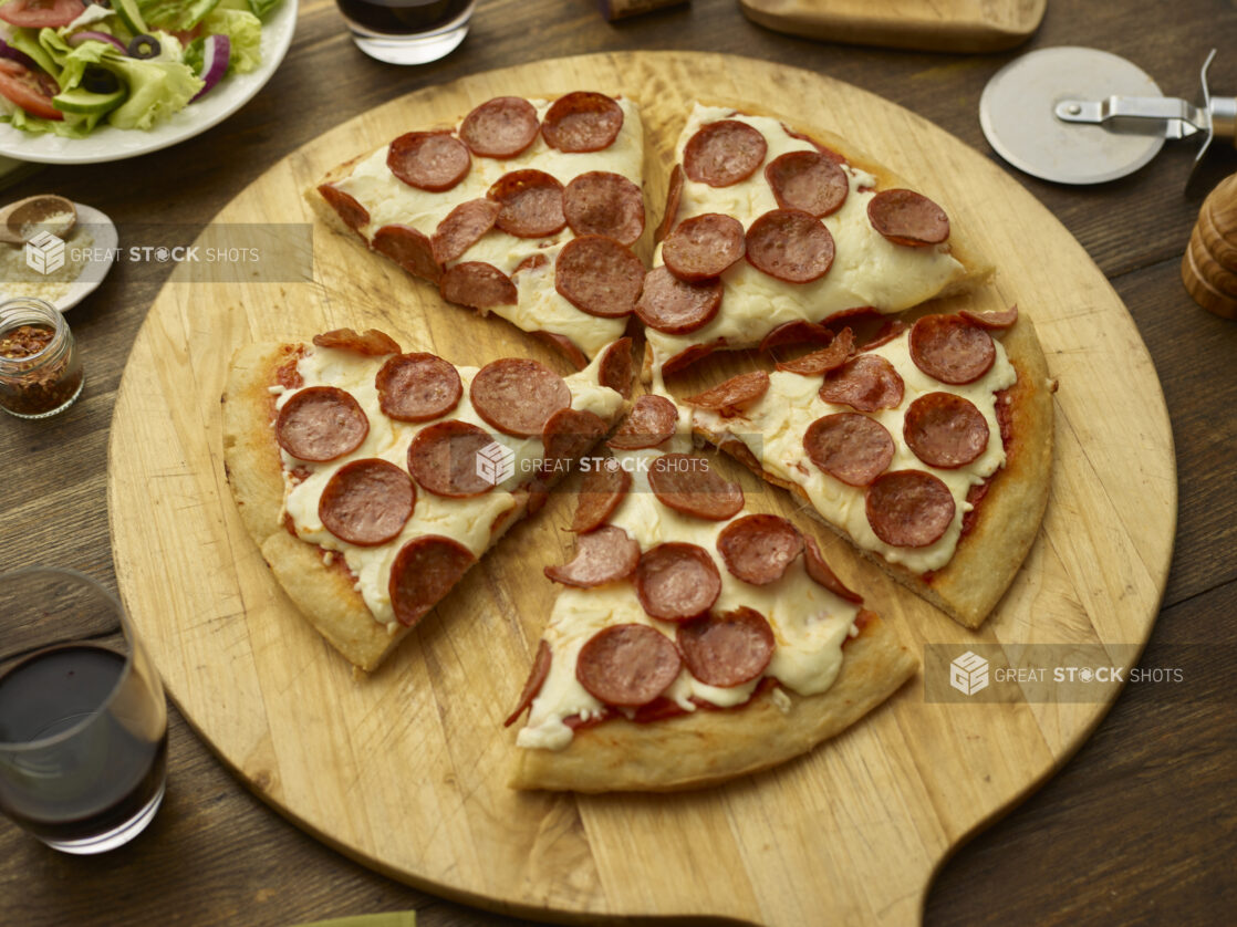 Sliced pepperoni pizza on a round wood pizza peel with pizza cutter, wooden tabletop