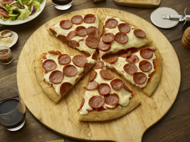 Sliced pepperoni pizza on a round wood pizza peel with pizza cutter, wooden tabletop