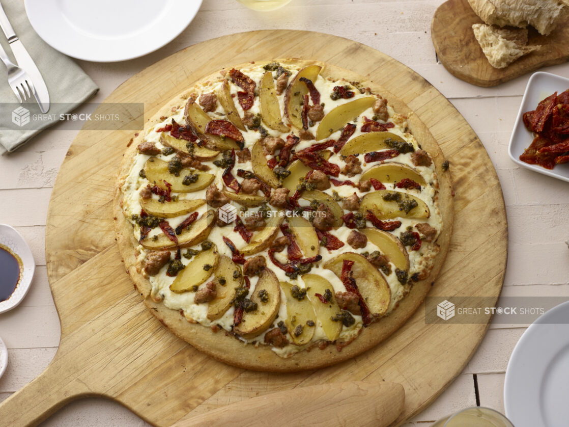 Whole pizza with potato wedges, sausage, sundried tomates, and pesto on a round wood pizza peel, overhead view