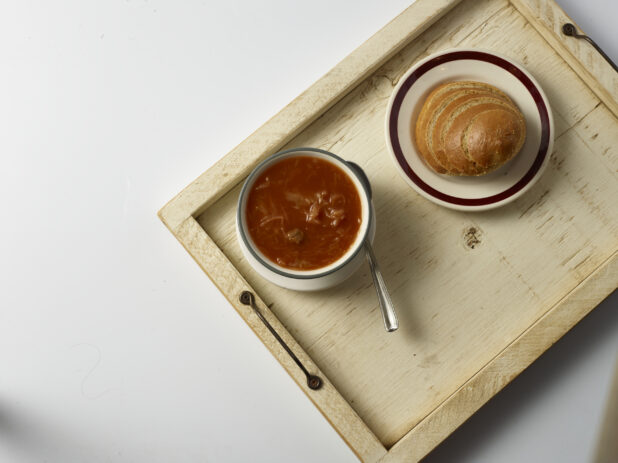 Overhead view of a bowl of cabbage soup with a plate of sliced light rye bread on a wood serving tray, white background