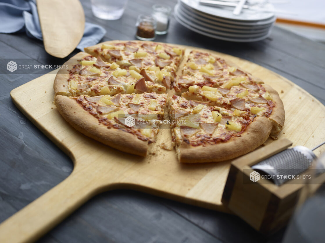 Sliced Hawaiian pizza on a wood pizza peel, dark grey background