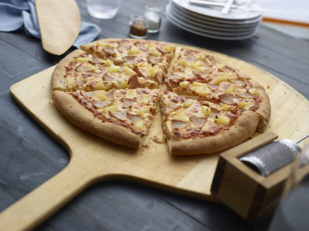 Sliced Hawaiian pizza on a wood pizza peel, dark grey background