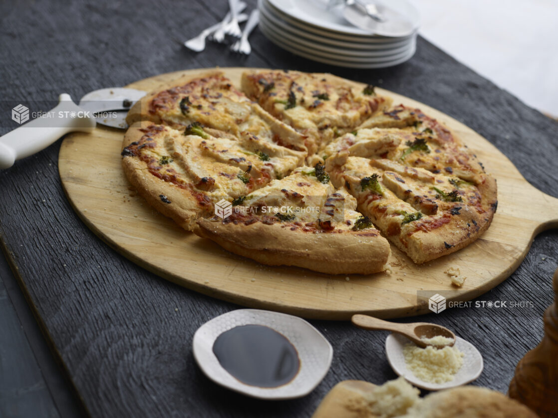 Sliced chicken, broccoli, and cheese pizza on a wood pizza peel with cutter, condiments and tableware surrounding, black background