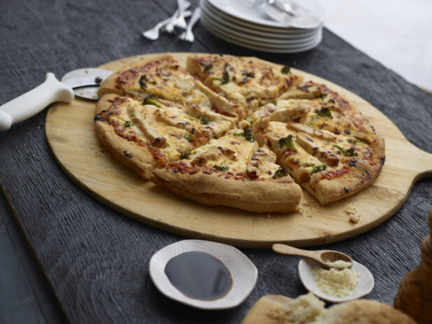 Sliced chicken, broccoli, and cheese pizza on a wood pizza peel with cutter, condiments and tableware surrounding, black background
