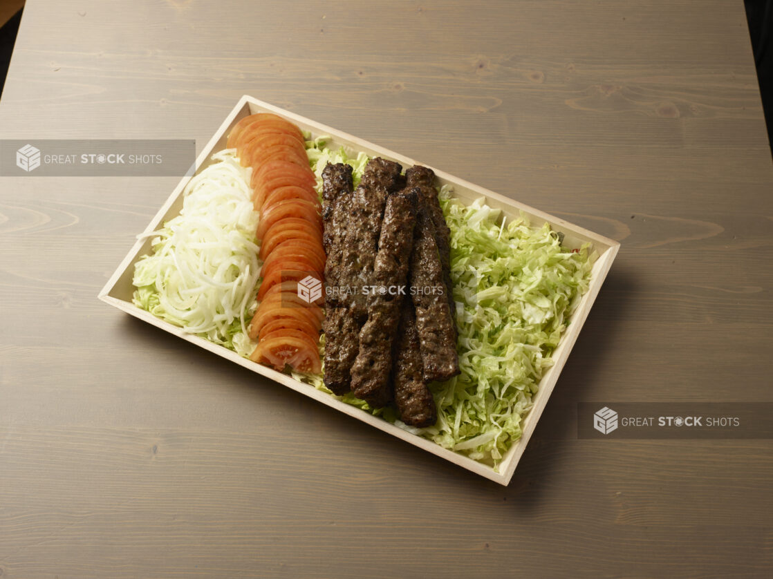 Overhead view of a wooden platter with sliced, onion, tomatoes and beef kebab on a bed of lettuce on a wooden background