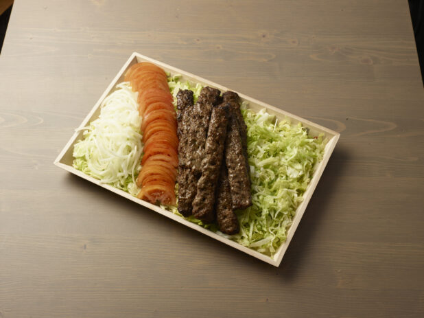 Overhead view of a wooden platter with sliced, onion, tomatoes and beef kebab on a bed of lettuce on a wooden background