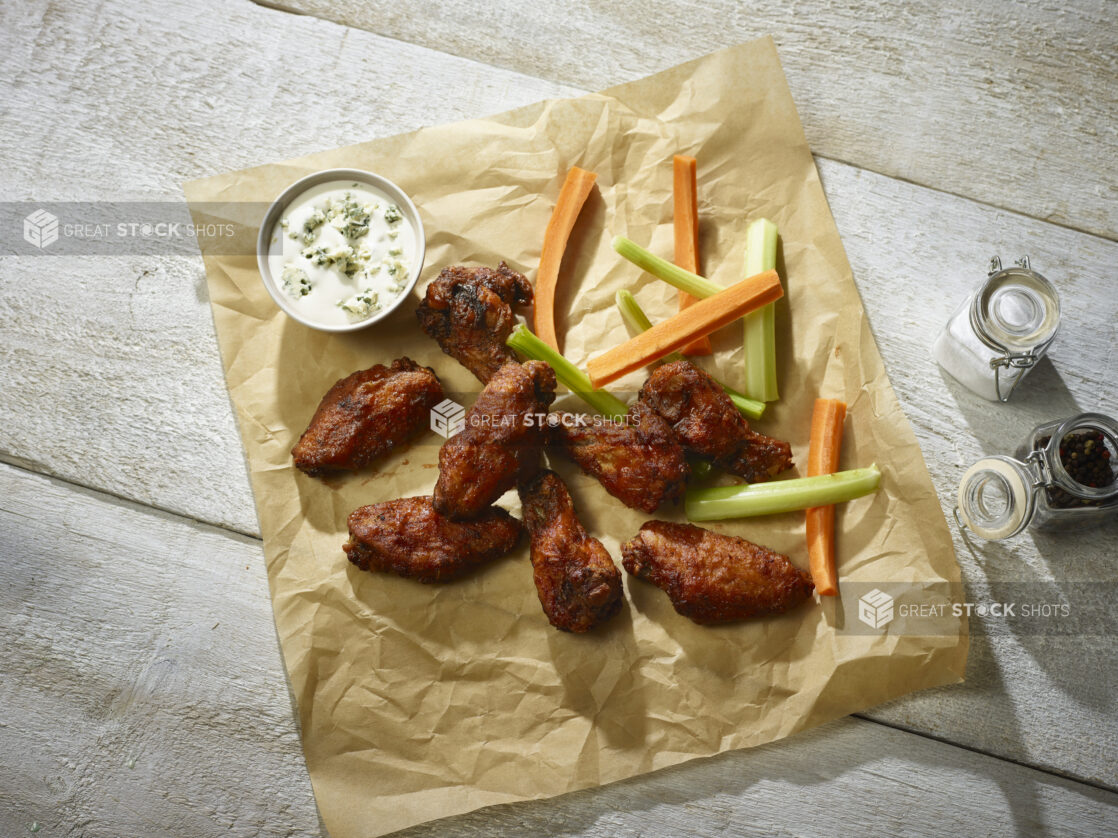 Barbecued chicken wings with celery and carrot sticks and a ramekin of blue cheese dip on parchment, salt and pepper beside, on a whitewashed wood background