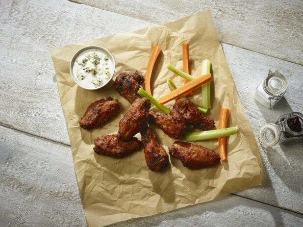 Barbecued chicken wings with celery and carrot sticks and a ramekin of blue cheese dip on parchment, salt and pepper beside, on a whitewashed wood background
