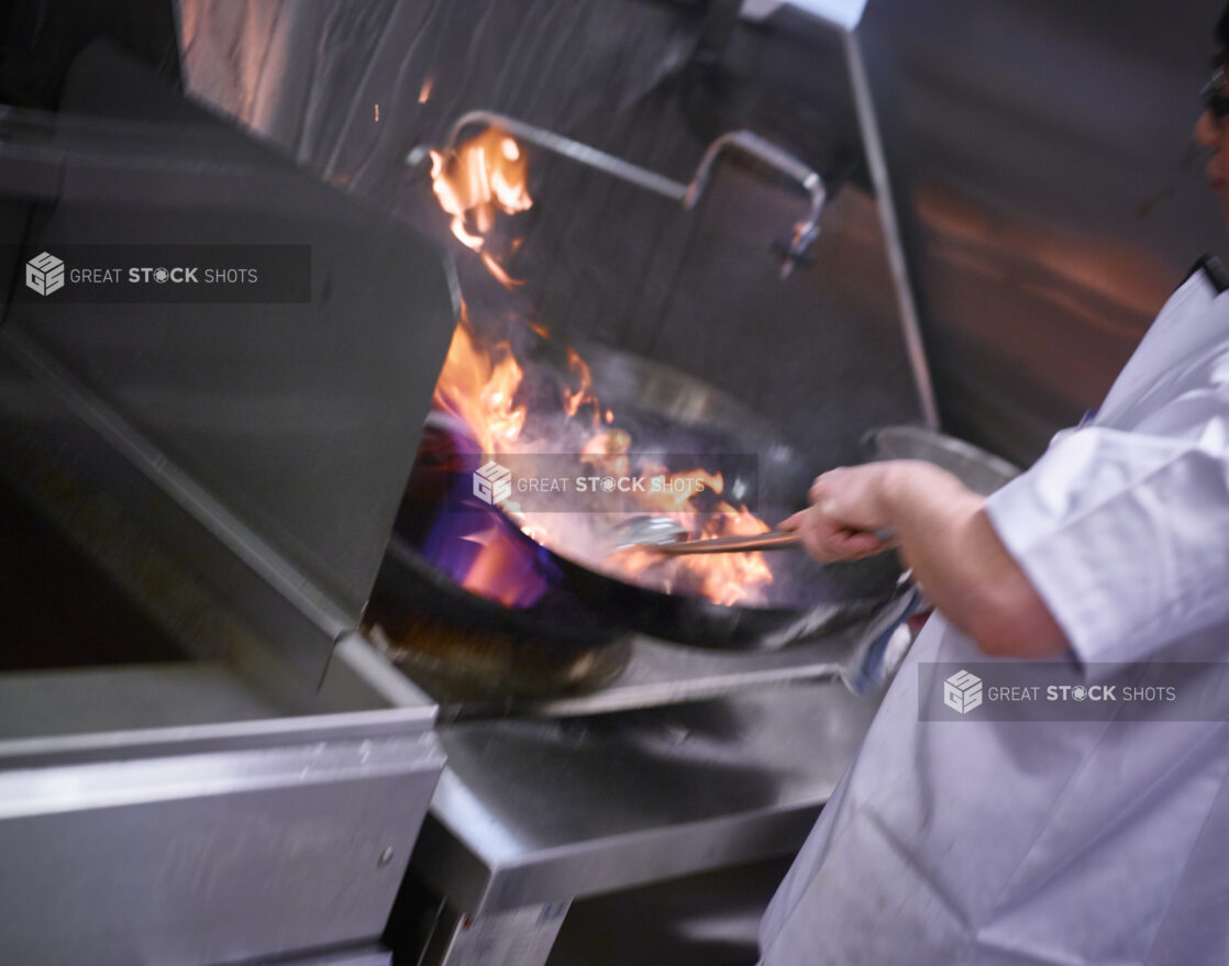 Chef stirring the contents of a flaming wok in a commercial kitchen setting