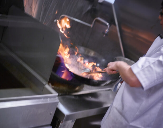 Chef stirring the contents of a flaming wok in a commercial kitchen setting