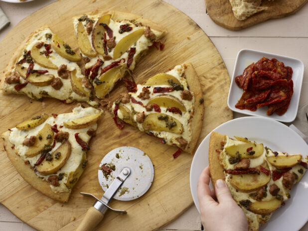 Sliced pizza with potato wedges, sausage, sundried tomates, and pesto on a round wood pizza peel, hand holding plate with pizza slice, overhead view