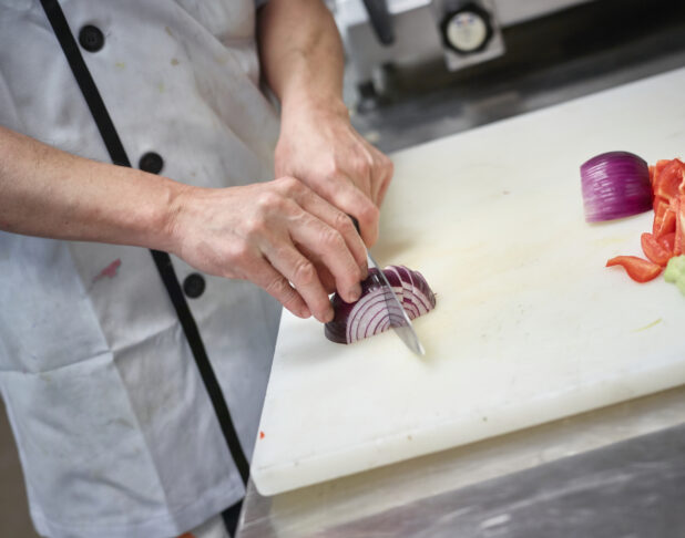 Prep cook cutting red onion on a white cutting board, kitchen setting