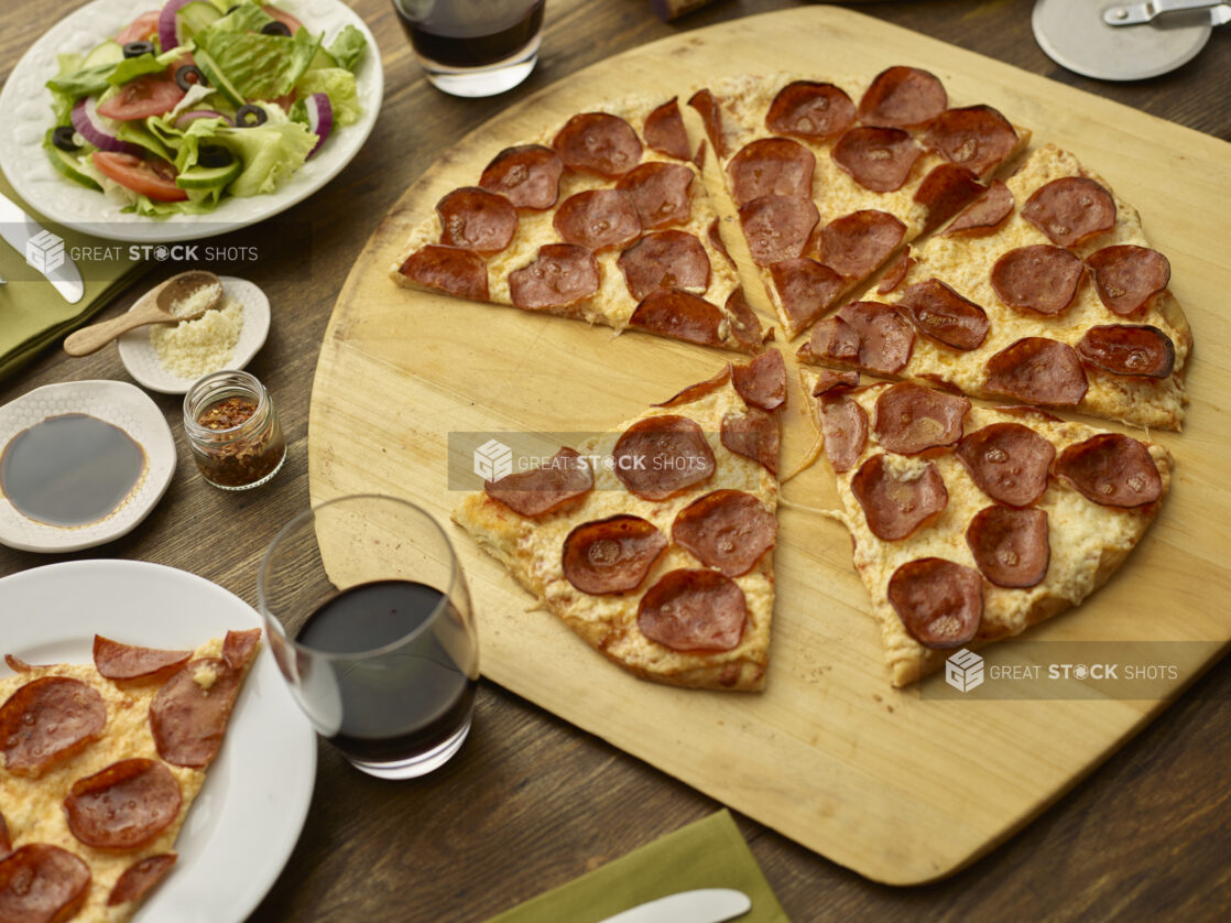 Sliced pepperoni pizza on a round wood pizza peel with pizza cutter, wooden tabletop, salad and glasses of wine surrounding