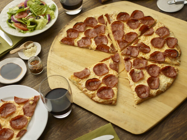 Sliced pepperoni pizza on a round wood pizza peel with pizza cutter, wooden tabletop, salad and glasses of wine surrounding